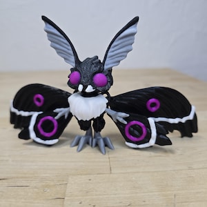 May include: A 3D-printed moth figurine with a detailed design. The moth features a black body, wings with white and purple accents, and large purple eyes. The wings are spread, and the figurine is posed on a wooden surface.