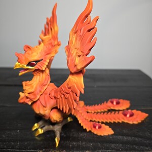 May include: A detailed, orange and yellow phoenix sculpture with outstretched wings and a long tail. The bird has a golden beak and talons, and red accents on its tail feathers. The sculpture is set against a dark background.