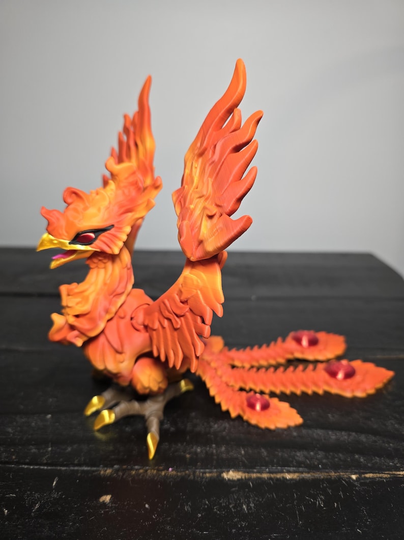 May include: A detailed, orange and yellow phoenix sculpture with outstretched wings and a long tail. The bird has a golden beak and talons, and red accents on its tail feathers. The sculpture is set against a dark background.