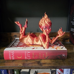 K&ouml;nnte beinhalten: Rote und orangefarbene Figur einer weiblichen Figur mit Flammen f&uuml;r Haare und H&auml;nde, die auf einem Buch mit dem Titel "Crescent City: House of Earth and Blood" von Sarah J. Maas liegt. Die Figur befindet sich auf einem Holzregal mit anderen B&uuml;chern.