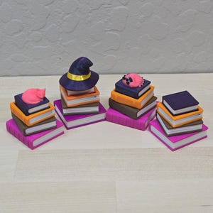 May include: Four stacks of miniature books in various colours including purple, orange, brown, and pink. One stack is topped with a purple witch hat with a yellow band, one with a pink cat, one with a pink creature, and one is plain.