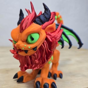 May include: A 3D-printed orange dragon figurine with red and black accents. The dragon has green eyes, black claws, and green spots. The wings are green and black. The figurine is posed on a wooden surface.