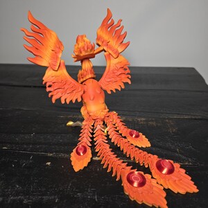 May include: A detailed orange and red phoenix sculpture with outstretched wings and flowing tail feathers. The sculpture features intricate detailing, with a fiery appearance. The tail feathers have red circular accents. The sculpture is set against a dark background.