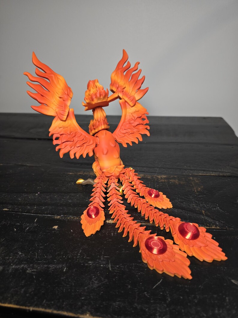 May include: A detailed orange and red phoenix sculpture with outstretched wings and flowing tail feathers. The sculpture features intricate detailing, with a fiery appearance. The tail feathers have red circular accents. The sculpture is set against a dark background.