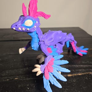 May include: A purple and blue articulated dinosaur toy with pink accents. The toy has a detailed design with feather-like structures and a stylized head. The dinosaur is posed on a dark surface, showcasing its features.