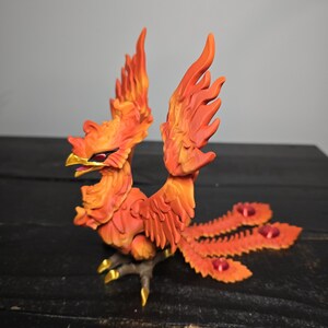 May include: A detailed sculpture of a phoenix in shades of orange and red, with gold accents on its beak and talons. The mythical bird has outstretched wings and a fanned tail, with red gems. The sculpture is on a dark surface.