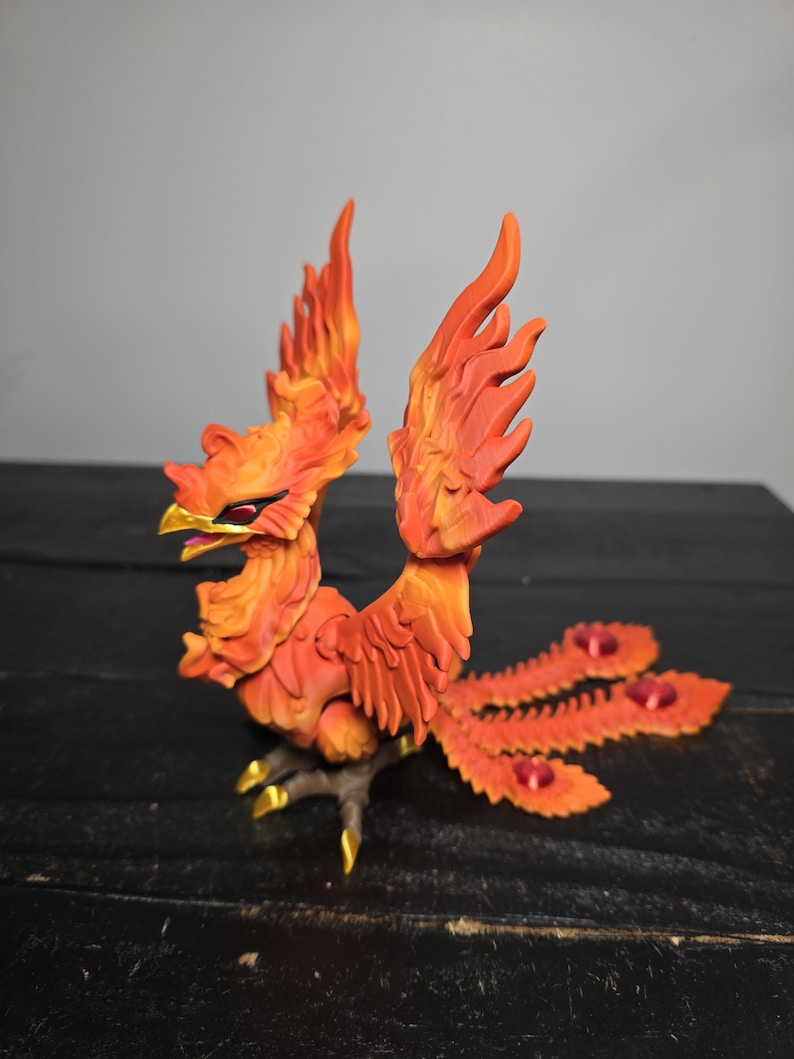 May include: A detailed sculpture of a phoenix in shades of orange and red, with gold accents on its beak and talons. The mythical bird has outstretched wings and a fanned tail, with red gems. The sculpture is on a dark surface.