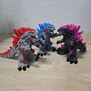 May include: Three Godzilla action figures in a row, each with a unique color scheme. The first is gray with red dorsal fins, the second is gray with blue fins, and the third is black with pink fins. All have detailed textures.