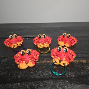 Peut inclure: Cinq porte-clés en forme de crabe rouge avec des pinces jaunes et des yeux noirs sont disposés sur une surface en bois sombre. Chaque crabe tient un anneau métallique dans ses pinces, avec des anneaux dorés, turquoise et violets. Accessoire amusant sur le thème de la plage.