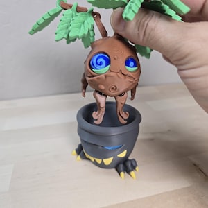 May include: A brown, plant-like creature figurine with large blue spiral eyes and green leaves on its head, suspended above a dark gray, pot-shaped base featuring a wide mouth with sharp yellow teeth and blue accents.