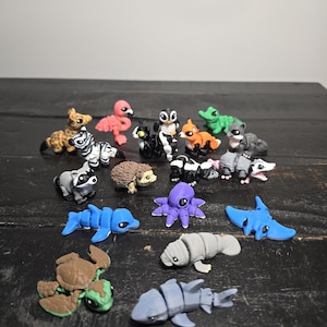May include: A selection of small, colourful animal figurines. The collection includes a zebra, flamingo, penguin, fox, alligator, raccoon, hedgehog, skunk, dolphin, octopus, manatee, shark, and turtle. The figurines are made of a smooth material and are on a dark wooden surface.
