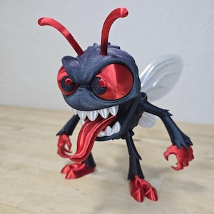 May include: A 3D-printed insect figurine with a menacing expression. The figure features a black body, red eyes, tongue, and feet, and white wings. The insect has a large, open mouth with sharp teeth and red antennae.