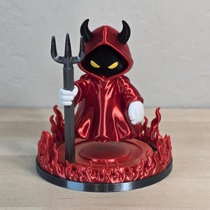 May include: A small figurine of a hooded character with black horns, yellow eyes, and a red robe, holding a black trident. The figure stands on a circular base surrounded by a ring of red flames, with white gloves visible on the character's hands.