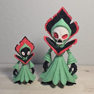 May include: Two stylized figures featuring a light green robe with a black and red collar and headpiece. The smaller figure has a black face with white eyes, while the larger figure has a white skull-like face with red eyes and an open mouth.