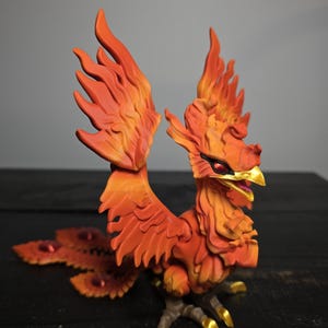 May include: A detailed figurine of a phoenix with fiery orange and yellow feathers. The mythical bird has a red eye, a golden beak, and golden talons. The wings are spread, and the tail features red accents.