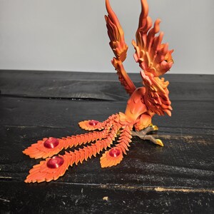 May include: A detailed orange and yellow phoenix sculpture with outstretched wings and a long, fanned tail. The sculpture features intricate feather detailing and red jewel accents. The bird is perched on a dark surface.