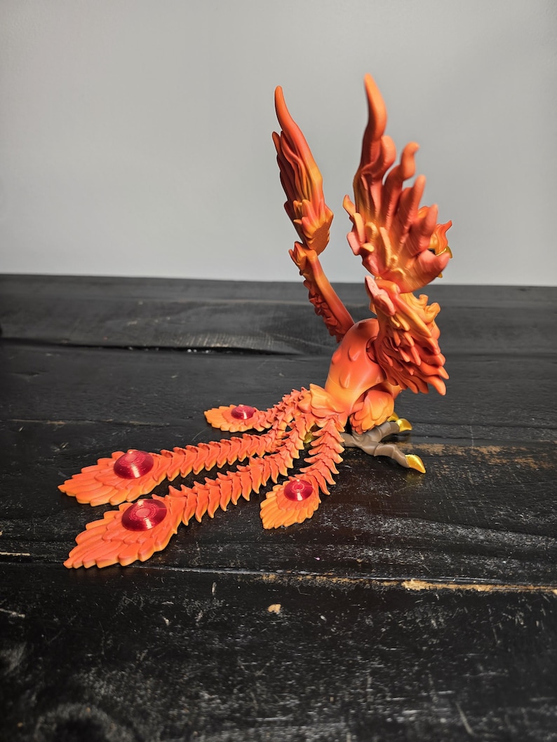 May include: A detailed orange and yellow phoenix sculpture with outstretched wings and a long, fanned tail. The sculpture features intricate feather detailing and red jewel accents. The bird is perched on a dark surface.