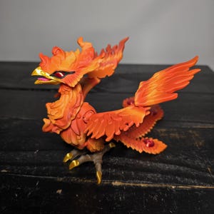 May include: A detailed figurine of a phoenix, predominantly orange with yellow and red accents. The mythical bird is depicted with outstretched wings and a fiery appearance. The figurine has a gold beak and claws, and is set against a dark background.