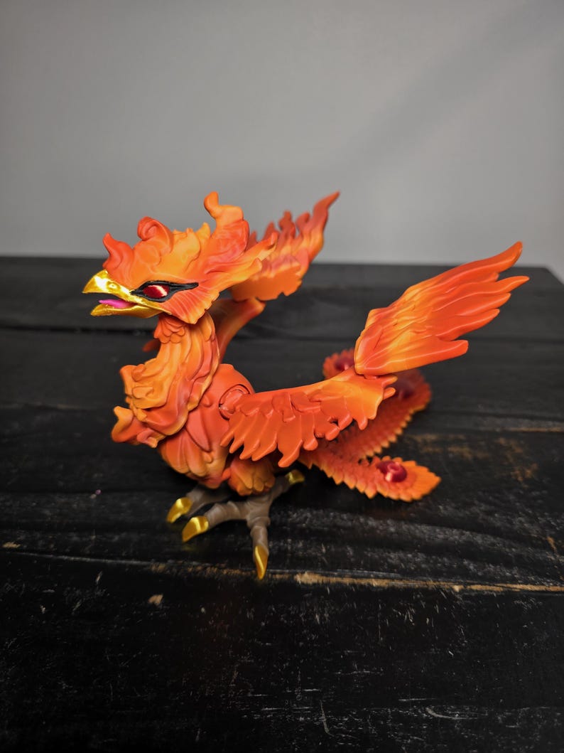 May include: A detailed figurine of a phoenix, predominantly orange with yellow and red accents. The mythical bird is depicted with outstretched wings and a fiery appearance. The figurine has a gold beak and claws, and is set against a dark background.
