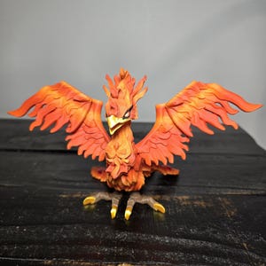 May include: A detailed figurine of a phoenix with outstretched wings. The bird is predominantly orange and red, with yellow accents on its talons and beak. The wings are spread wide, showcasing intricate feather details.