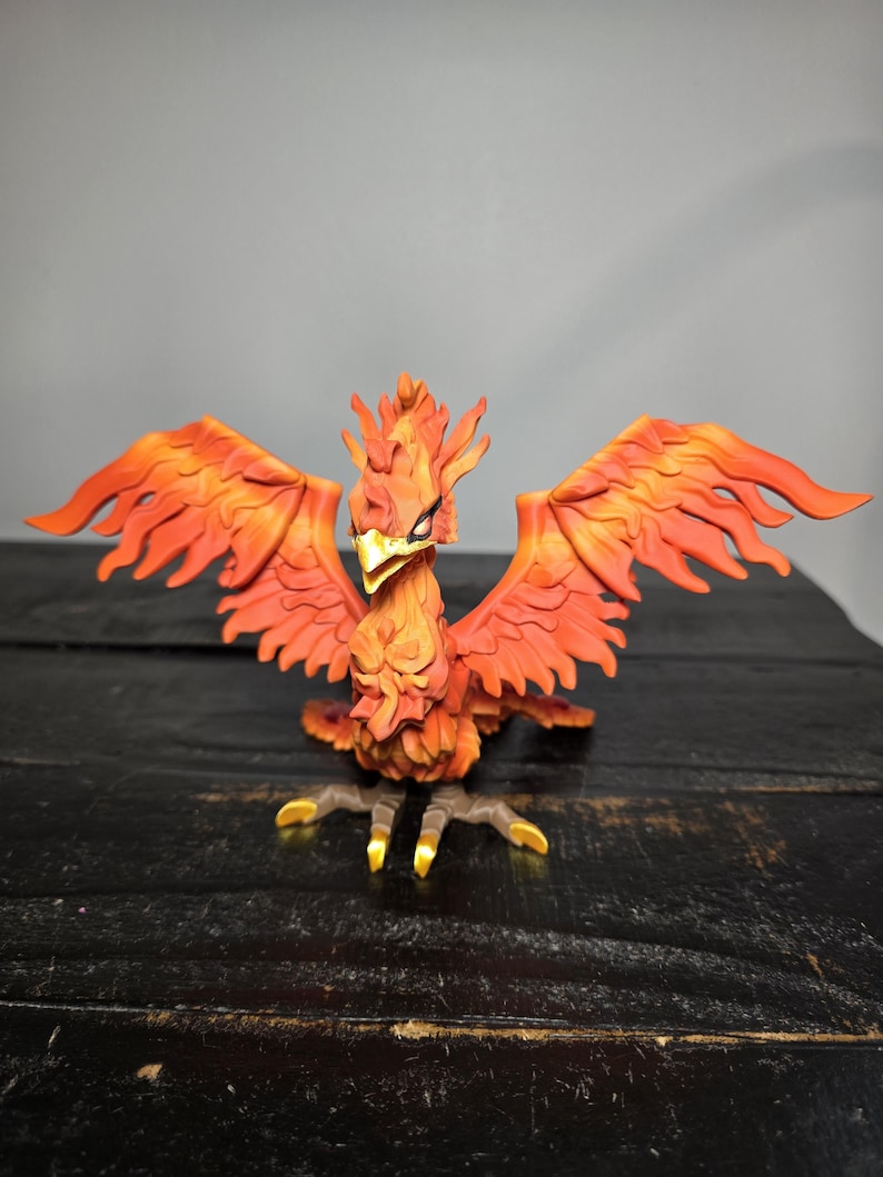 May include: A detailed figurine of a phoenix with outstretched wings. The bird is predominantly orange and red, with yellow accents on its talons and beak. The wings are spread wide, showcasing intricate feather details.