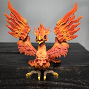 May include: A detailed figurine of a phoenix with outstretched wings. The bird is predominantly orange and red, with intricate feather detailing. Its beak is open, and its talons are gold. The figurine is set against a dark background.