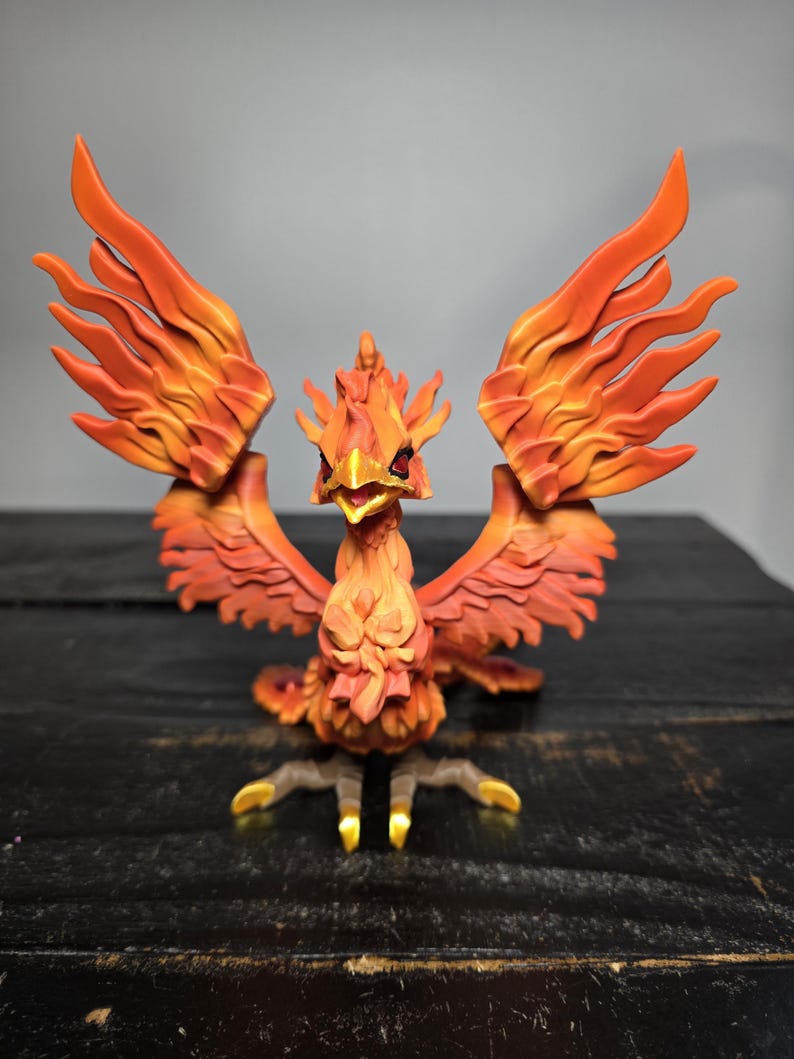 May include: A detailed figurine of a phoenix with outstretched wings. The bird is predominantly orange and red, with intricate feather detailing. Its beak is open, and its talons are gold. The figurine is set against a dark background.