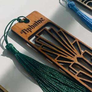 May include: A rectangular wooden bookmark featuring a laser-cut geometric sunburst pattern and the name Tiphaine engraved at the top. A dark green thread tassel is attached through a circular hole at the top edge of the wooden bookmark.