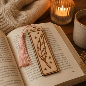 May include: A rectangular light-colored wooden bookmark featuring a laser-engraved feather design, four small stars, and the name AURÉLIE at the top. A soft pink tassel is attached to the top hole of the bookmark, which rests on an open book page.