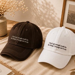 Op de afbeelding: Twee baseball caps, één bruin en één wit, worden getoond. Elke cap heeft de geborduurde tekst: "I don't get enough credit for the things I manage not to say". Ze liggen op een beige oppervlak.