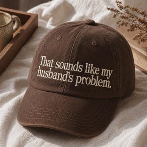 Könnte beinhalten: Braune Baseballkappe mit dem gestickten Text "That sounds like my husband's problem." Die Kappe hat einen gebogenen Schirm und eine runde Oberseite. Der Text ist in weißer, serifenloser Schrift.
