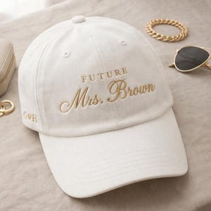 Puede incluir: Gorra de béisbol blanca con la inscripción bordada en hilo dorado "FUTURE Mrs. Brown". La gorra tiene una visera curva y un botón en la parte superior. Las iniciales "G H" están bordadas en el lateral. Gafas de sol y una pulsera son visibles en el fondo.