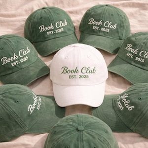 Może przedstawiać: Kolekcja zielonych i białych czapek z daszkiem. Każda czapka ma napis "Book Club EST. 2025" w kontrastowym kolorze. Czapki są ułożone w okrąg, z białą czapką pośrodku.
