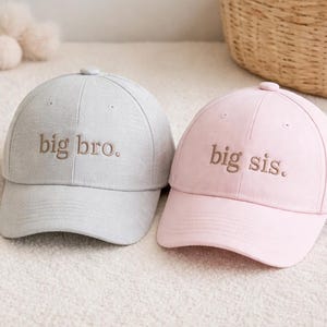 Puede incluir: Dos gorras de béisbol, una gris claro y otra rosa. La gorra gris tiene bordado "big bro." en hilo marrón, mientras que la gorra rosa dice "big sis.". Ambas gorras tienen una visera curva y un botón en la parte superior.