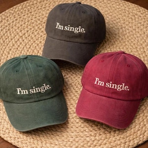 Puede incluir: Tres gorras de béisbol en diferentes colores: gris oscuro, verde y burdeos. Cada gorra tiene el texto bordado "I'm single". Las gorras están dispuestas sobre una superficie beige tejida.