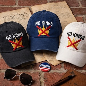 Puede incluir: Tres gorras de béisbol en negro, azul marino y blanco, cada una bordada con "NO KINGS" sobre una corona tachada. También se ve un pin de "FREEDOM" y gafas de sol. Las gorras descansan sobre una superficie de ladrillo con documentos históricos.