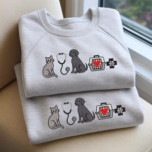 Puede incluir: Dos sudaderas de color gris claro apiladas, cada una con un diseño bordado. El diseño incluye un gato gris, un estetoscopio, un perro negro, un transportín para mascotas con un corazón rojo y una cruz con huella de pata. Las sudaderas están dobladas.