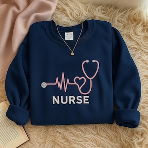 May include: Navy blue sweatshirt with a pink stethoscope design and the word "NURSE" in white. A gold necklace with a small pendant is visible. The sweatshirt is made of a soft material.