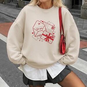 May include: Beige sweatshirt with a red graphic of an envelope with "love you" text, a gift box, and postcards. The design includes hearts. A red purse hangs from the shoulder.