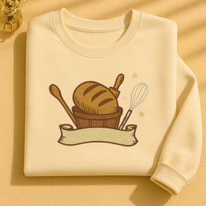 Könnte beinhalten: Cremefarbenes Sweatshirt mit einem gestickten Design von Backutensilien und einem Brotlaib. Das Design umfasst einen Holzlöffel, eine Teigrolle, einen Schneebesen und ein Banner. Das Sweatshirt ist ein Rundhals-Stil.