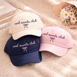May include: Three baseball caps in cream, navy blue, and pink. Each cap has the embroidered text "cool aunts club" in a matching color, with a bow graphic. The caps are arranged on a soft, textured surface.