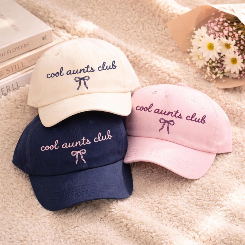 May include: Three baseball caps in cream, navy blue, and pink. Each cap has the embroidered text "cool aunts club" in a matching color, with a bow graphic. The caps are arranged on a soft, textured surface.