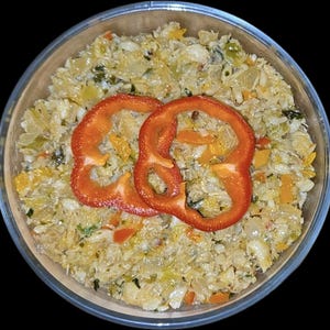 May include: A clear glass bowl filled with a colorful dish. The dish features a base of finely chopped vegetables, with visible pieces of yellow, green, and orange. Two vibrant red bell pepper rings are placed on top, creating a striking contrast.