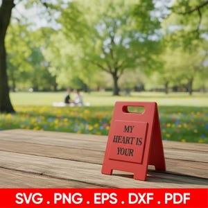 Op de afbeelding: Een rood A-frame bord met de tekst "MY HEART IS YOUR" staat op een houten oppervlak. Het bord staat voor een vage achtergrond van een park met bomen en bloemen. De onderkant van de afbeelding heeft de tekst "SVG . PNG . EPS . DXF . PDF".