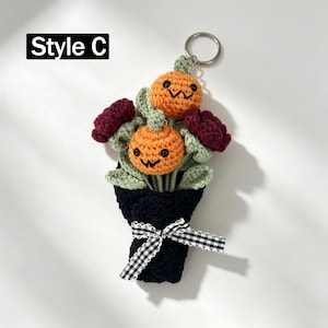 May include: A crochet keychain labeled Style C featuring a bouquet of two orange pumpkins with black embroidered faces, two dark red flowers, and light green leaves. The bouquet is wrapped in black yarn and tied with a black and white gingham ribbon.