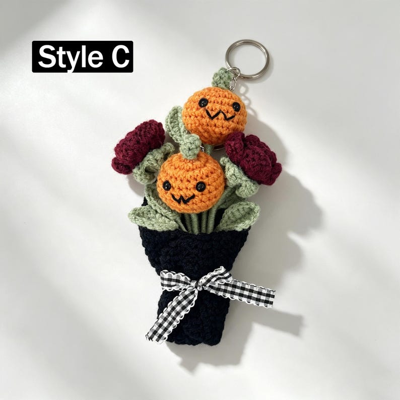 May include: A crochet keychain labeled Style C featuring a bouquet of two orange pumpkins with black embroidered faces, two dark red flowers, and light green leaves. The bouquet is wrapped in black yarn and tied with a black and white gingham ribbon.