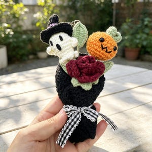 May include: A small crochet bouquet featuring a white ghost wearing a black witch hat, a bright orange pumpkin with a face, and a deep red rose. The bouquet is wrapped in black yarn and tied with a black and white gingham ribbon bow.