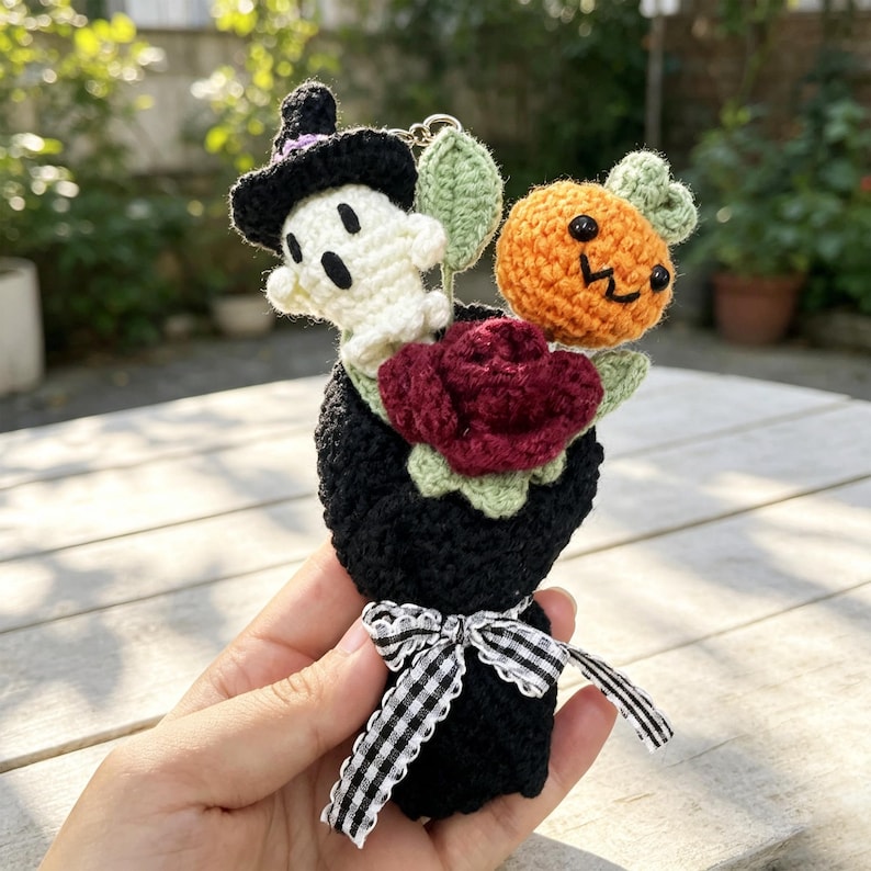 May include: A small crochet bouquet featuring a white ghost wearing a black witch hat, a bright orange pumpkin with a face, and a deep red rose. The bouquet is wrapped in black yarn and tied with a black and white gingham ribbon bow.