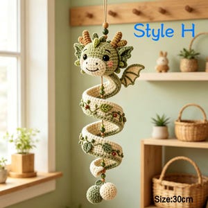 K&ouml;nnte beinhalten: Ein h&auml;ngendes H&auml;kel-Drachenornament in Salbeigr&uuml;n und Creme mit einem spiralf&ouml;rmigen K&ouml;rper, der mit kleinen gr&uuml;nen Bl&auml;ttern und braunen Perlen verziert ist. Der Drache hat ein l&auml;chelndes Gesicht, kleine Fl&uuml;gel und eine L&auml;nge von 30 cm. Text Style H.
