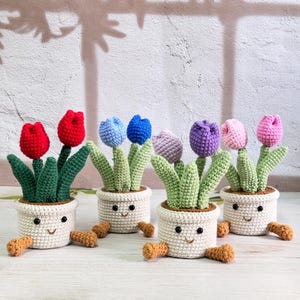 Puede incluir: Cinco plantas de tulipanes en maceta de ganchillo con caras sonrientes. Los tulipanes son rojos, azules, morados, rosas y lavanda. Las macetas son blancas con patas marrones.